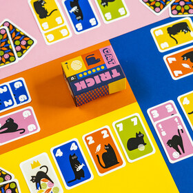 Draufsicht auf ein buntes Stichkartenspiel namens „Cat Trick". Auf einem vierfarbigen Hintergrund aus Pink, Orange, Gelb und Blau sind zahlreiche Spielkarten ausgebreitet. Die Karten zeigen schwarze Katzensilhouetten auf farbigen Hintergründen (Pink, Blau, Gelb, Orange, Grün) mit weißen Zahlen in den Ecken. In der Bildmitte steht die kleine quadratische Spielschachtel mit dem Aufdruck „Cat Trick". Links liegen einige Karten mit dekorativem Rückenmuster aus Blättern, Wollknäueln und Fischen.