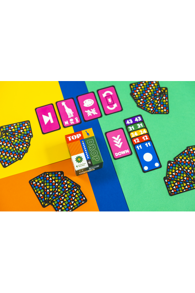 Lifestyle-Flatlay auf einem vierfarbig geteilten Hintergrund (Gelb, Orange, Blau, Grün). Die Spielschachtel steht in der Bildmitte. Drumherum sind Karten ausgebreitet: oben pinke Sonderkarten mit Symbolen wie Pfeil, Giraffe, Farbpalette und Drehpfeil; rechts eine Reihe aufgedeckter Zahlenkarten in absteigender Folge (43, 31, 24, 12, 11) in Lila, Grün, Gelb, Rot und Blau sowie eine blaue Karte mit weißem Kreis; an den Rändern mehrere Stapel verdeckter Karten mit buntem Symbolmuster.