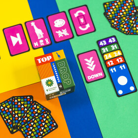 Lifestyle-Flatlay auf einem vierfarbig geteilten Hintergrund (Gelb, Orange, Blau, Grün). Die Spielschachtel steht in der Bildmitte. Drumherum sind Karten ausgebreitet: oben pinke Sonderkarten mit Symbolen wie Pfeil, Giraffe, Farbpalette und Drehpfeil; rechts eine Reihe aufgedeckter Zahlenkarten in absteigender Folge (43, 31, 24, 12, 11) in Lila, Grün, Gelb, Rot und Blau sowie eine blaue Karte mit weißem Kreis; an den Rändern mehrere Stapel verdeckter Karten mit buntem Symbolmuster.