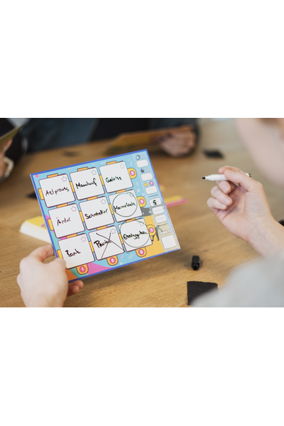 Zwei Hände halten ein buntes Spieltableau aus dem Brettspiel „Dito!" in die Kamera. Auf dem türkis-gelb-pinken Tableau sind neun beschreibbare Felder in einem 3\times3-Raster zu sehen, handschriftlich befüllt mit Wörtern wie „Arztpraxis", „Maulwurf", „Gebiss", „Ämter", „Schmetterling", „Marmelade", „Bank", „Pavisc" und „Erdnüsse". Zwei Felder sind mit einem Kreis markiert, eines mit einem Kreuz durchgestrichen. Im Hintergrund sitzt eine weitere Person am Holztisch; eine Hand hält einen abwischbaren Stift.