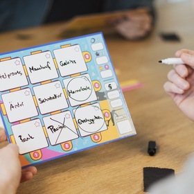 Zwei Hände halten ein buntes Spieltableau aus dem Brettspiel „Dito!" in die Kamera. Auf dem türkis-gelb-pinken Tableau sind neun beschreibbare Felder in einem 3\times3-Raster zu sehen, handschriftlich befüllt mit Wörtern wie „Arztpraxis", „Maulwurf", „Gebiss", „Ämter", „Schmetterling", „Marmelade", „Bank", „Pavisc" und „Erdnüsse". Zwei Felder sind mit einem Kreis markiert, eines mit einem Kreuz durchgestrichen. Im Hintergrund sitzt eine weitere Person am Holztisch; eine Hand hält einen abwischbaren Stift.