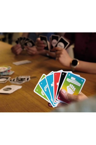 Eine Hand hält einen Fächer aus sechs bunten Frantic-Spielkarten, darunter eine Counterattack-Karte mit Faust-Symbol. Im Hintergrund ein Holztisch, auf dem weitere Karten liegen; zwei weitere Personen halten ebenfalls Karten in den Händen.