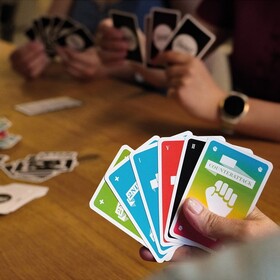 Eine Hand hält einen Fächer aus sechs bunten Frantic-Spielkarten, darunter eine Counterattack-Karte mit Faust-Symbol. Im Hintergrund ein Holztisch, auf dem weitere Karten liegen; zwei weitere Personen halten ebenfalls Karten in den Händen.