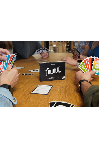 Vier Personen sitzen an einem Holztisch und spielen Frantic. Jede Person hält einen Fächer bunter Spielkarten. In der Tischmitte steht die schwarze Spielschachtel, daneben liegt eine Regelkarte zu den einfarbigen Spezialkarten sowie einzelne aufgedeckte Karten. Die Szene vermittelt eine lebhafte Spielrunde.