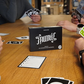 Vier Personen sitzen an einem Holztisch und spielen Frantic. Jede Person hält einen Fächer bunter Spielkarten. In der Tischmitte steht die schwarze Spielschachtel, daneben liegt eine Regelkarte zu den einfarbigen Spezialkarten sowie einzelne aufgedeckte Karten. Die Szene vermittelt eine lebhafte Spielrunde.