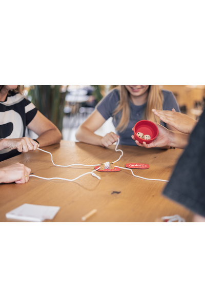 Nahaufnahme eines laufenden QORK-Spiels auf einem Holztisch. In der Bildmitte eine rote Spielmatte mit mehreren Korken, von denen weiße Schnüre sternförmig zu den Spielerinnen und Spielern führen. Rechts hält eine Person den roten Becher mit zwei Würfeln darin. Links ziehen Hände an den Schnüren. Im Hintergrund sind Oberkörper von zwei Personen zu sehen, unscharf. Die Szene vermittelt den Moment kurz vor oder nach dem Würfeln.