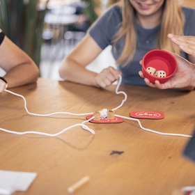 Nahaufnahme eines laufenden QORK-Spiels auf einem Holztisch. In der Bildmitte eine rote Spielmatte mit mehreren Korken, von denen weiße Schnüre sternförmig zu den Spielerinnen und Spielern führen. Rechts hält eine Person den roten Becher mit zwei Würfeln darin. Links ziehen Hände an den Schnüren. Im Hintergrund sind Oberkörper von zwei Personen zu sehen, unscharf. Die Szene vermittelt den Moment kurz vor oder nach dem Würfeln.