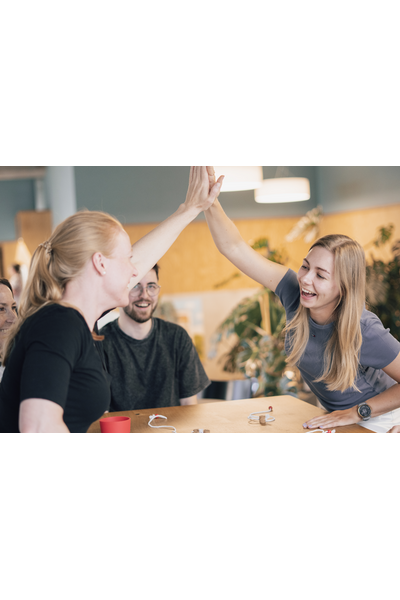 Vier Personen sitzen lachend um einen Holztisch und spielen gemeinsam QORK. Im Mittelpunkt eine Frau mittleren Alters mit schwarzem Oberteil, die sich über den Tisch beugt und einen roten Becher festhält. Ihr gegenüber ein junger Mann mit Brille und Bart, der ebenfalls lacht und eine Schnur hält. Links und rechts weitere Spielerinnen, von denen nur Schultern und Arme zu sehen sind. Auf dem Tisch liegen weiße Spielmatten und Schnüre. Im Hintergrund helle Fenster und eine große Zimmerpflanze.
