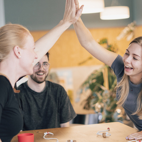Vier Personen sitzen lachend um einen Holztisch und spielen gemeinsam QORK. Im Mittelpunkt eine Frau mittleren Alters mit schwarzem Oberteil, die sich über den Tisch beugt und einen roten Becher festhält. Ihr gegenüber ein junger Mann mit Brille und Bart, der ebenfalls lacht und eine Schnur hält. Links und rechts weitere Spielerinnen, von denen nur Schultern und Arme zu sehen sind. Auf dem Tisch liegen weiße Spielmatten und Schnüre. Im Hintergrund helle Fenster und eine große Zimmerpflanze.