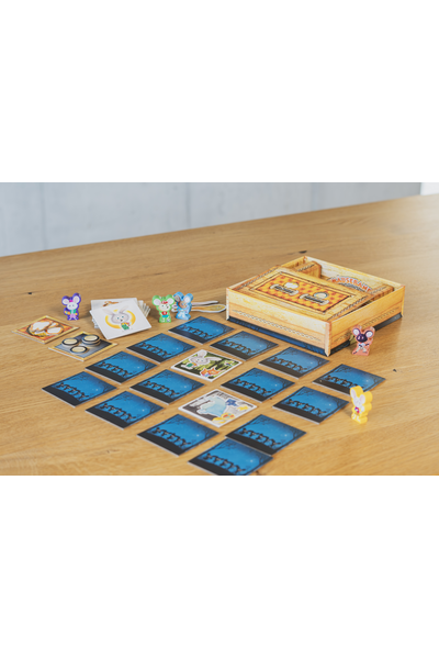 Auf einem hellen Holztisch liegt das aufgebaute Kinderspiel „Die Mäusebande". Im Zentrum sind etwa 20 quadratische Plättchen mit blauem Nacht-Motiv (Silhouetten von Mäusen vor Bäumen und Sternenhimmel) in einem Raster ausgelegt, zwei davon sind umgedreht und zeigen bunte Tierfiguren. Oben rechts steht die goldbraune Holzspielschachtel. Verteilt auf dem Tisch stehen fünf kleine bunte Holzmäuse-Figuren (lila, grün, blau, lila-rosa, gelb). Links liegen Stapel mit Karten und goldig gerahmten Zahn-Plättchen sowie zwei Münzen.