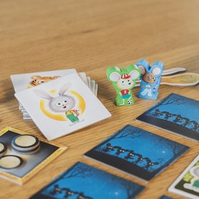 Nahaufnahme des Spielmaterials auf einem Holztisch. Im Vordergrund liegen mehrere blaue Nacht-Plättchen und ein aufgedecktes Plättchen mit einem Hasen in einem grünen Wald. In der Bildmitte liegt ein Stapel weißer Karten, die oberste zeigt einen grauen Hasen vor gelbem Hintergrund. Dahinter stehen zwei Holzmäuse-Figuren (grün und blau) sowie ein hölzerner Schlüssel. Links steht eine lila Maus-Figur, daneben goldig gerahmte Zahn-Plättchen mit Münzen. Am rechten Rand ist die Spielbox angeschnitten zu sehen.